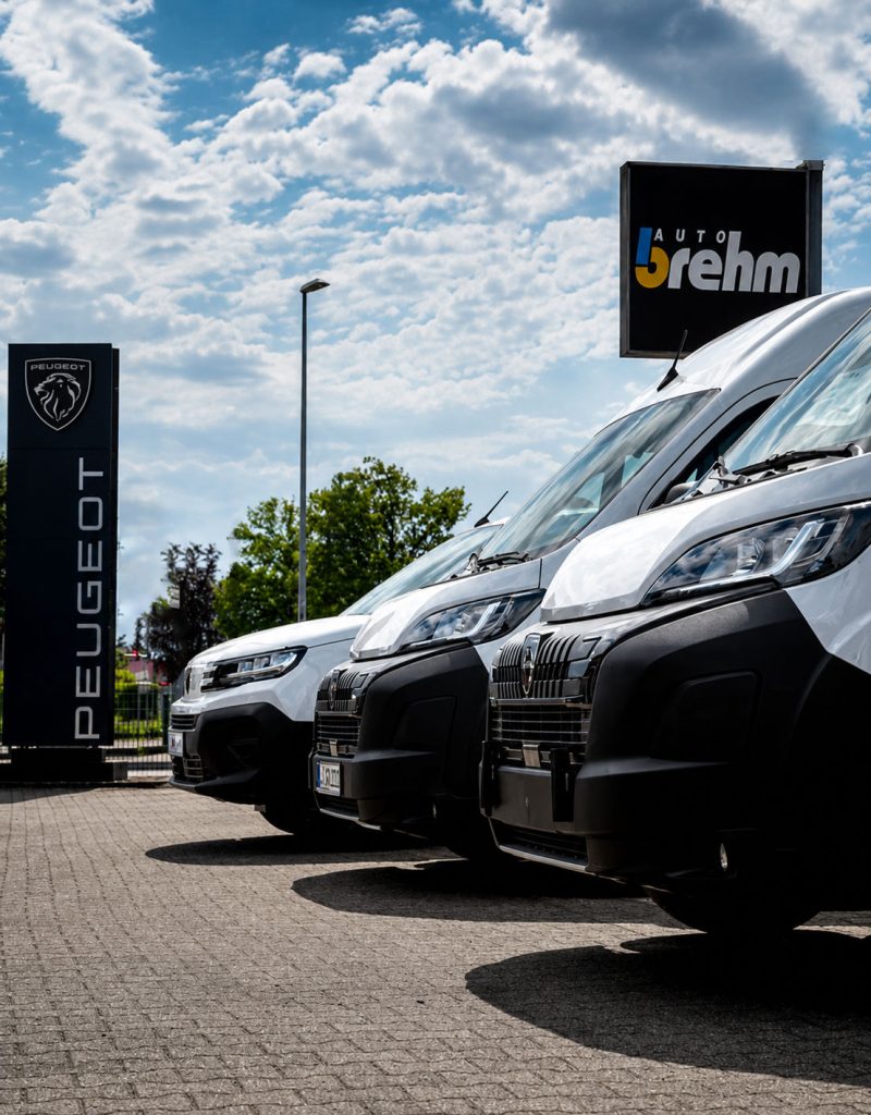 Mehrere weiße Peugeot-Fahrzeuge stehen auf dem Gelände des Autohauses Brehm neben Markenschildern unter blauem Himmel.